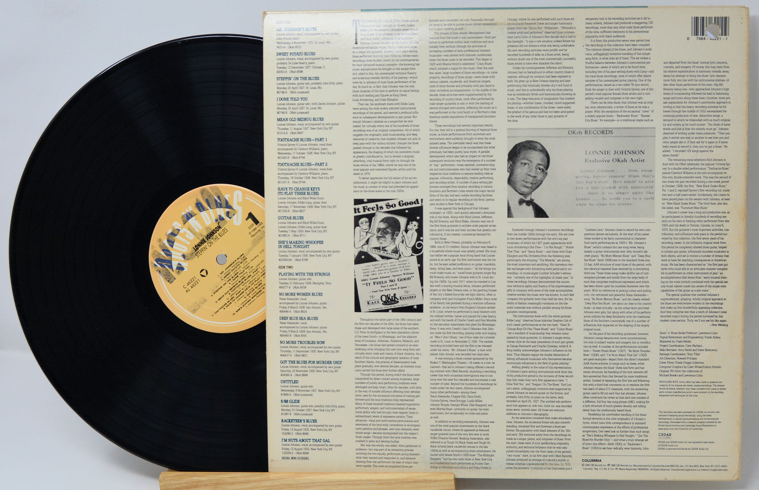A vinyl record partially pulled from its back cover sleeve, displayed on a small wooden stand against a light gray background. The black record has a tan label, and the sleeve is cream-colored with dense text and a small black-and-white portrait.