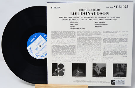 A vinyl record partially pulled from its printed album sleeve, showing the black disc with a blue-and-white label beside the white cover art for Lou Donaldson's 'The Time Is Right'.