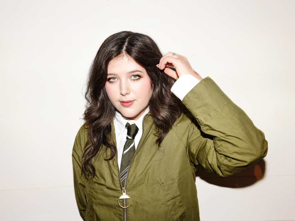 Lucy Dacus wearing a green jacket over a white shirt with a tie against a plain background.
