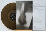 A vinyl record partially pulled from its sleeve, displayed on a small wooden stand against a white background. The record is translucent gold, and the sleeve features a black-and-white abstract photo on the front and a tracklist on the back.
