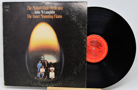Vinyl record album for 'The Inner Mounting Flame' by The Mahavishith Orchestra and John McLaughlin, showing the black cover with an orange-yellow oval graphic and the black record partially pulled out, revealing a red-orange center label.