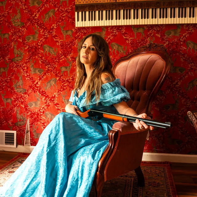 A woman in a long blue dress sits in a brown armchair holding a shotgun, set against a red patterned wallpaper background with a piano keyboard visible at the top.