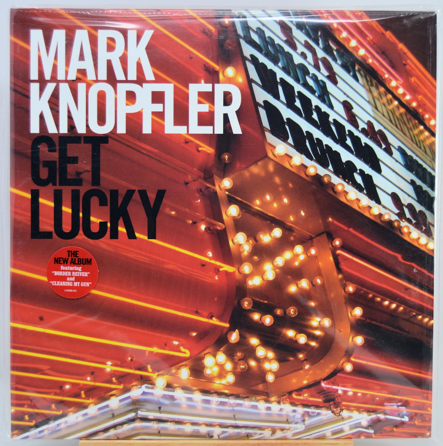 Mark Knopfler's 'Get Lucky' vinyl record album cover, featuring a vibrant orange and red marquee scene with bright light bulbs and bold white and black text.
