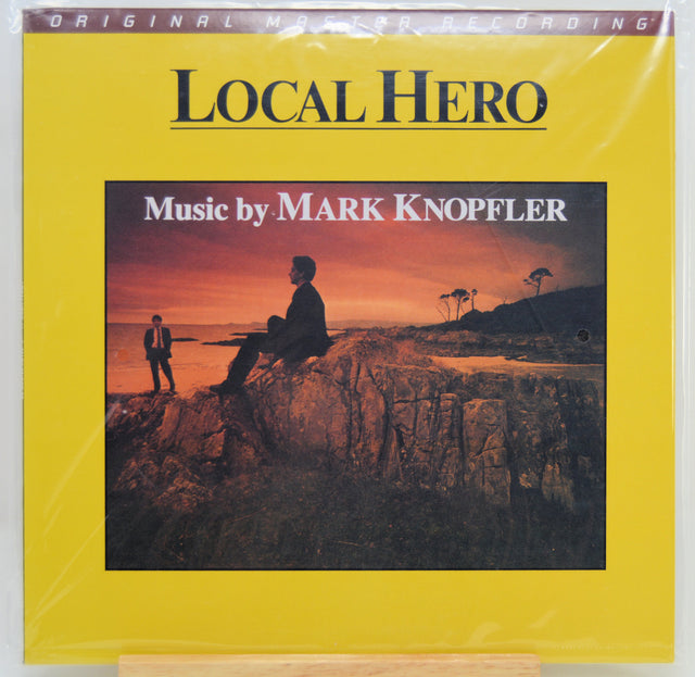Local Hero original music recording album cover by Mark Knopfler, featuring a bright yellow border and a central photograph of a coastal sunset scene with silhouetted figures on rocks.