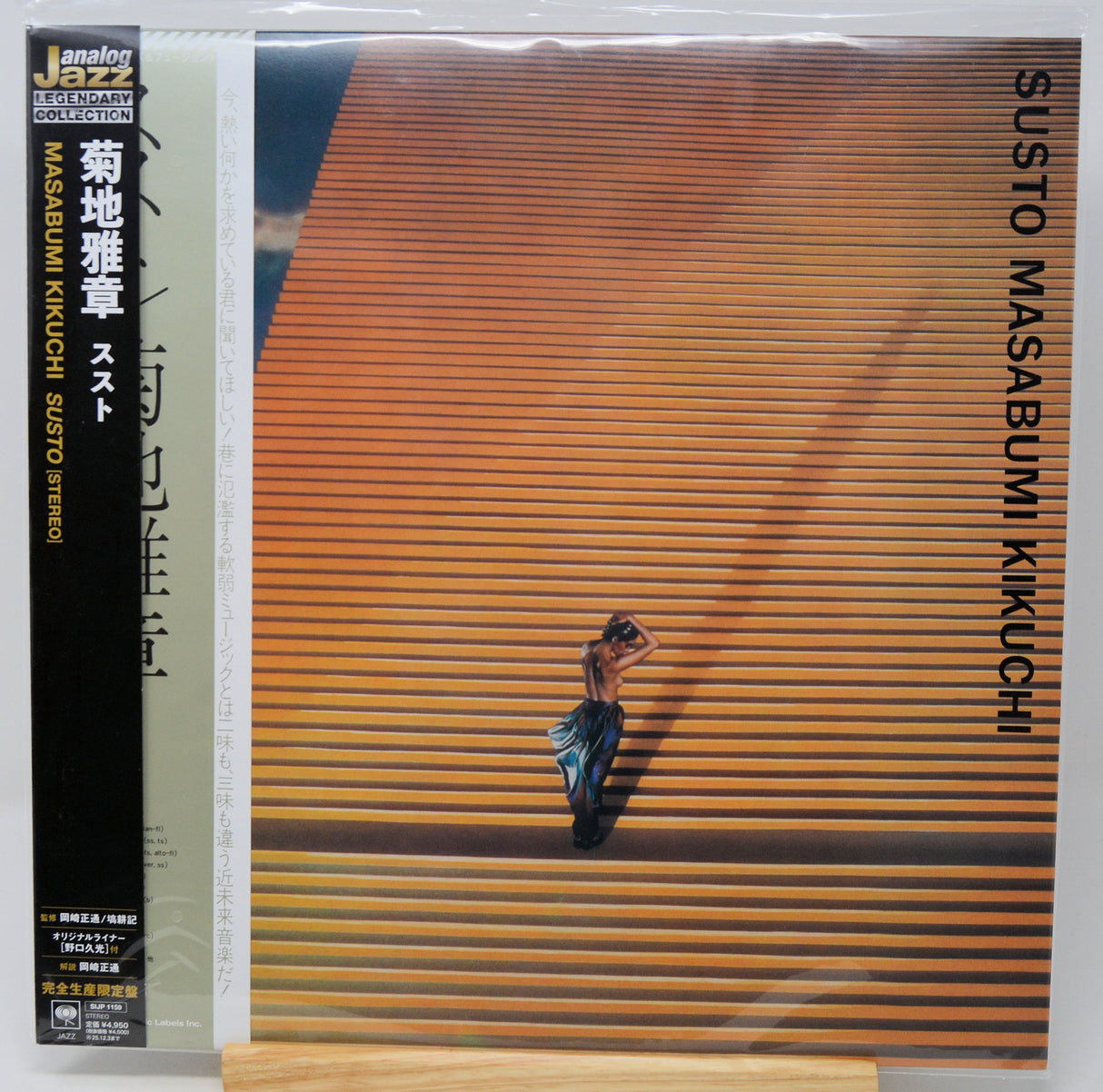 Vinyl record album cover for Susuto Masabumi Kikuchi's 'Susto' on a wooden stand. The cover features a striking orange and brown striped background with a small figure in the center, a black spine band, and a clear plastic shrink wrap.