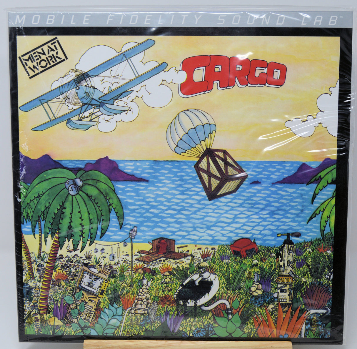 A square vinyl record sleeve for the album 'Cargo' by Metal Work, featuring a highly detailed, colorful cartoon illustration of a beach scene with a biplane, hot air balloon, palm trees, and a black-and-white cat.