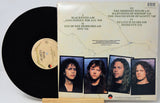 A vinyl record partially pulled from its sleeve, showing the black disc and the printed album cover. The cover includes the tracklist and four portrait photos of band members.