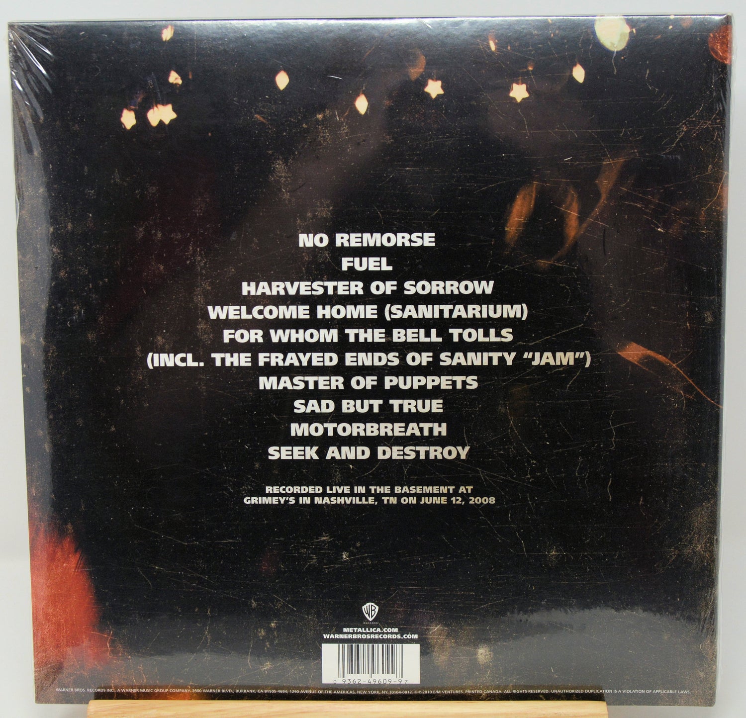 The back cover of a vinyl record album wrapped in plastic, showing tracklist text on a black background with small gold star-like graphics and a small red area in the lower left corner.