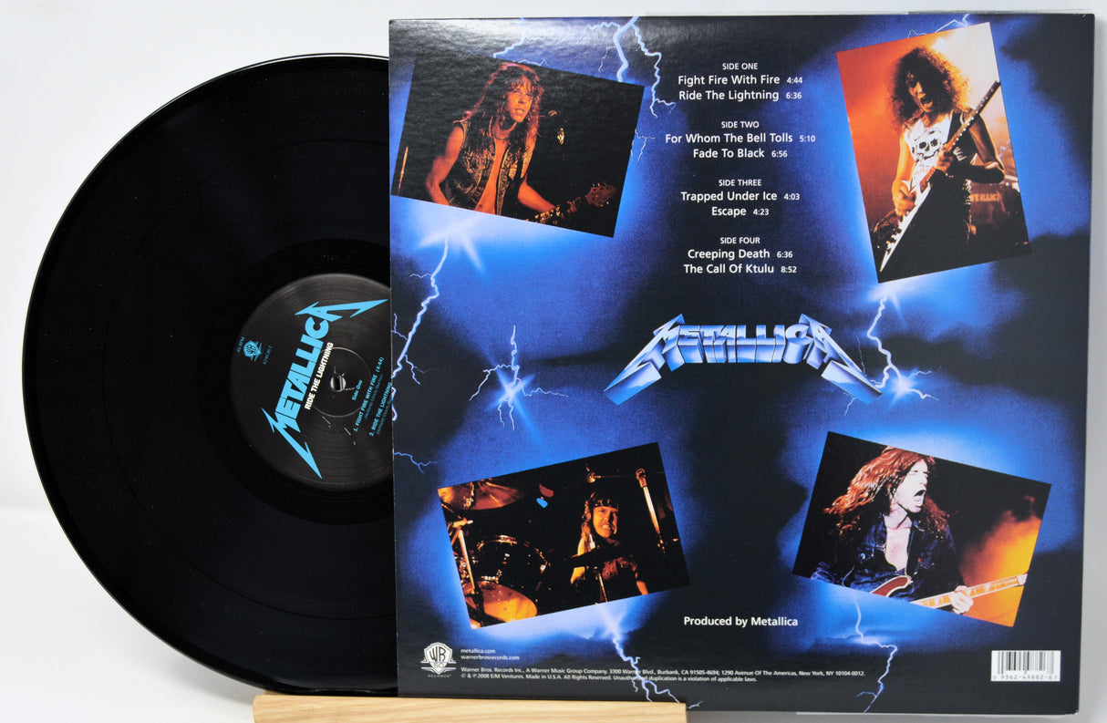 Metallica vinyl record shown with the back of the album cover visible beside the black record. The cover features blue lightning artwork, the Metallica logo, and four band photos on a white background.