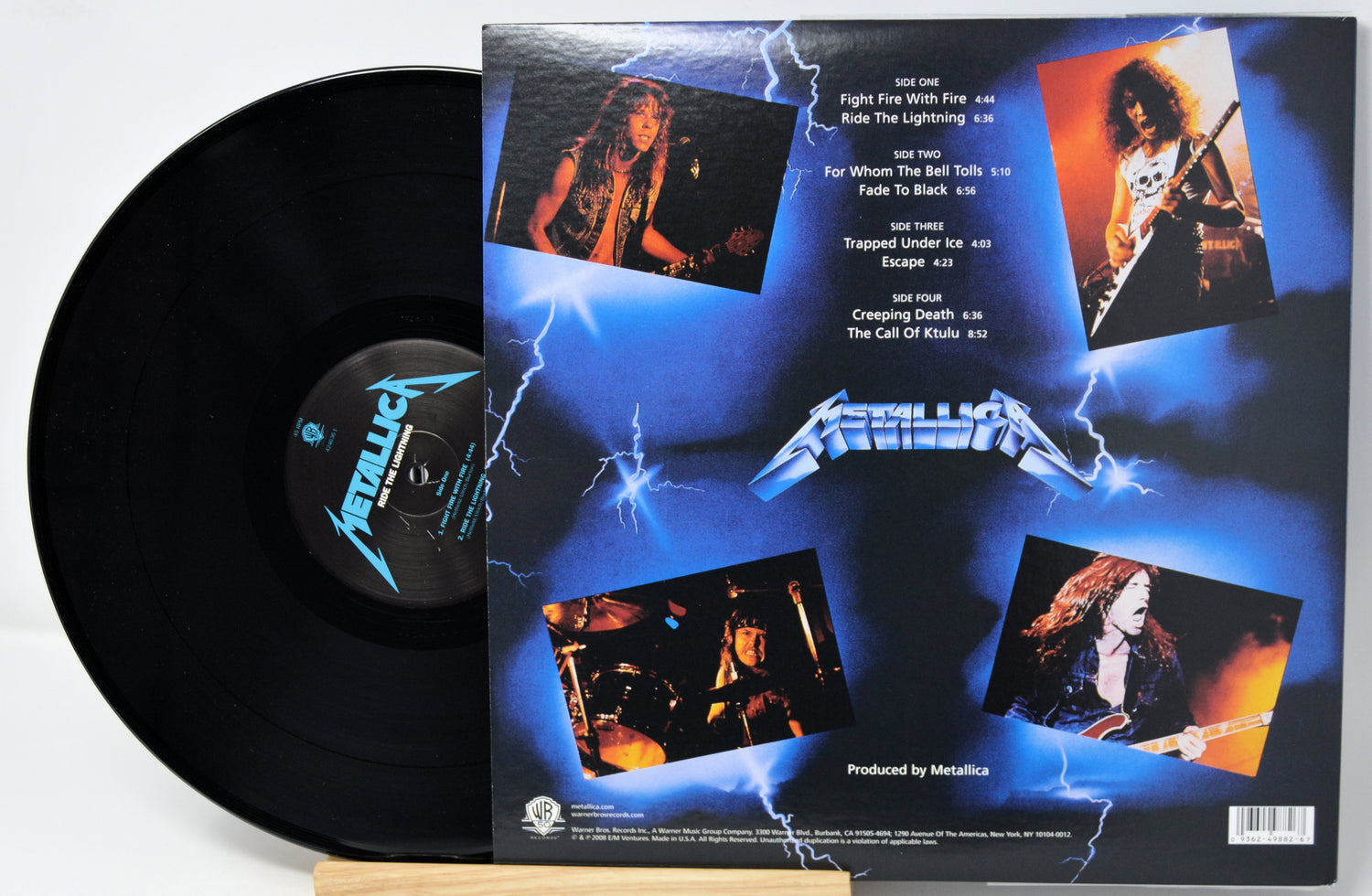 Metallica vinyl record shown with the back of the album cover visible beside the black record. The cover features blue lightning artwork, the Metallica logo, and four band photos on a white background.