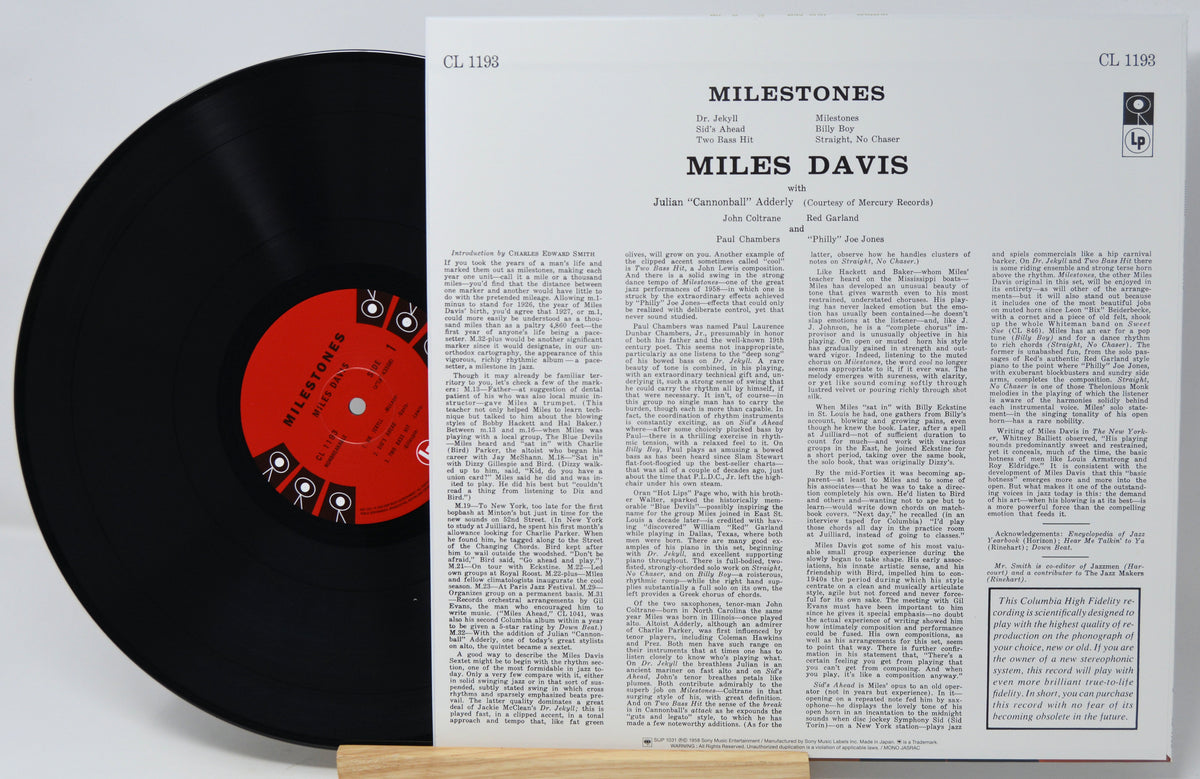 A vinyl record partially pulled from its printed back cover, showing the black record with a red center label beside the white printed album insert for Miles Davis' 'Milestones'.