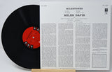 A vinyl record partially pulled from its printed back cover, showing the black record with a red center label beside the white printed album insert for Miles Davis' 'Milestones'.