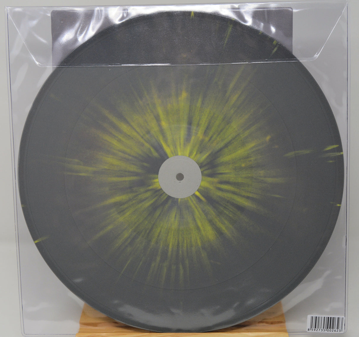 A square vinyl record in a clear plastic protective sleeve, featuring a dark gray/black disc with a bright yellow-green radial burst pattern from the center, displayed on a small wooden stand.