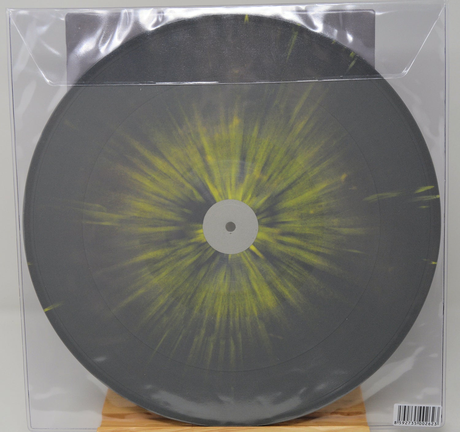 A square vinyl record in a clear plastic protective sleeve, featuring a dark gray/black disc with a bright yellow-green radial burst pattern from the center, displayed on a small wooden stand.