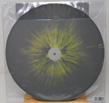 A square vinyl record in a clear plastic protective sleeve, featuring a dark gray/black disc with a bright yellow-green radial burst pattern from the center, displayed on a small wooden stand.