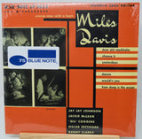 Miles Davis '75 Blue Note' vinyl record album cover, sealed in plastic shrink wrap, displayed on a wooden stand against a white background.