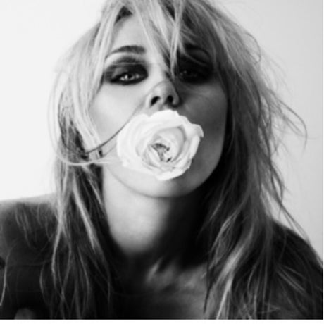 A black and white close-up portrait of a woman holding a white rose in her mouth, with long tousled hair and dark eye makeup against a plain light background.