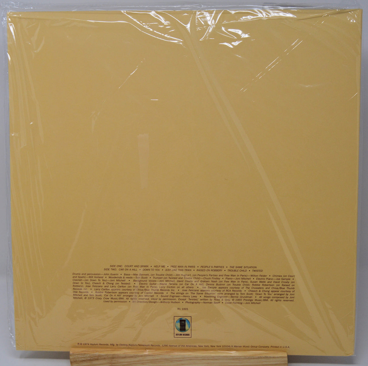 A vinyl record sleeve or album cover in clear plastic shrink-wrap, photographed flat against a light gray background. The cover is a warm mustard/golden yellow with small black text and a small square logo near the bottom center.