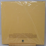 A vinyl record sleeve or album cover in clear plastic shrink-wrap, photographed flat against a light gray background. The cover is a warm mustard/golden yellow with small black text and a small square logo near the bottom center.