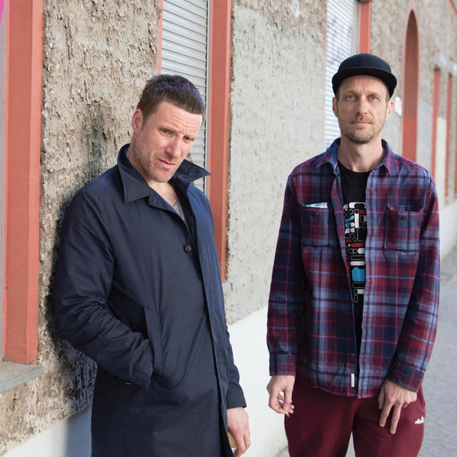 Two men in urban streetwear posing against a textured outdoor wall, one in a navy overcoat and one in a plaid shirt and cap