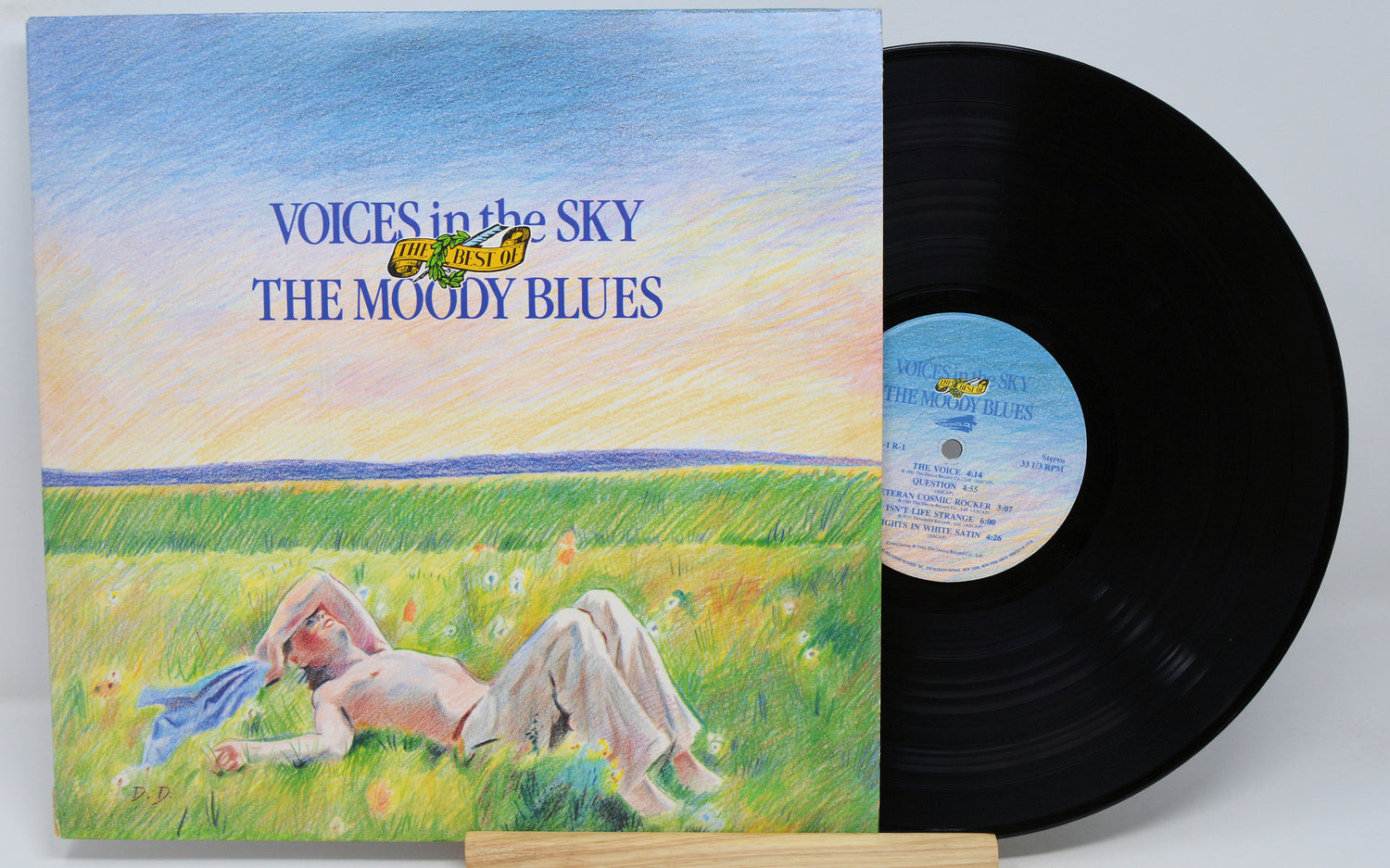 Used vinyl record of Moody Blues - Voices In The Sky with a record cover featuring an illustration of a person relaxing on grass under a blue sky, next to a vinyl record.