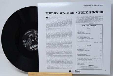 A black vinyl record partially visible behind an open printed album insert for Muddy Waters, folk singer, with track listing and printed text on the white page.
