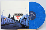 Mudhoney 'Tomorrow Hit Today' vinyl record album with the cover sleeve on the left and a translucent blue marbled record partially visible on the right, displayed on a small wooden stand against a light gray background.