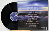 A vinyl record partially pulled from its sleeve, showing the black record on the left and the album cover on the right. The cover features a scenic landscape with a sunset sky and white handwritten-style text listing song titles.