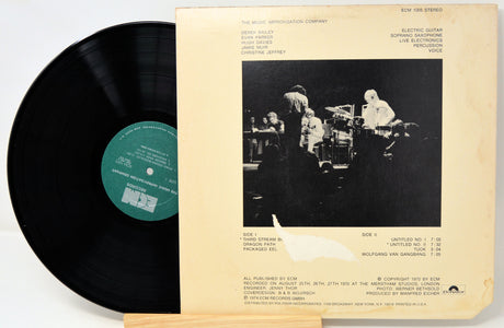 A vinyl record partially pulled from its cream-colored sleeve, showing the black record with a green label on the left and the back cover artwork on the right, photographed on a light wood surface.