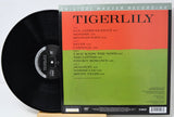 Vinyl record album 'Tigerlily' showing the black LP disc partially pulled from its sleeve, with the cover featuring a bright orange-red upper half and a lime green lower half, plus tracklist text on the front.
