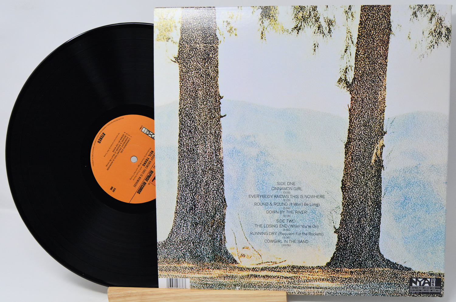 A vinyl record partially pulled from its sleeve, showing the black record with an orange label next to the album cover featuring two large tree trunks against a pale sky.