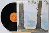 A vinyl record partially pulled from its sleeve, showing the black record with an orange label next to the album cover featuring two large tree trunks against a pale sky.