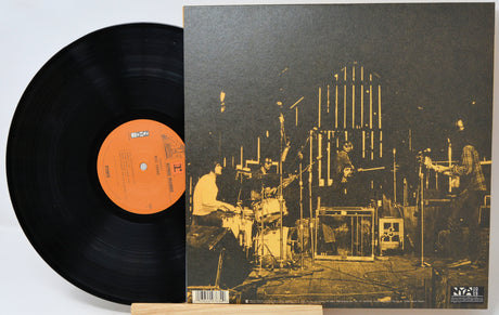 A vinyl record partially pulled from its sleeve, showing the black record with an orange center label and the album cover featuring a sepia-toned concert scene.