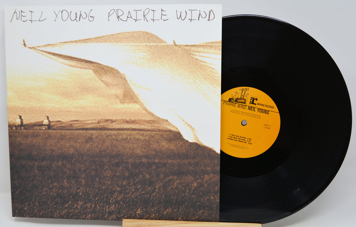 Neil Young's 'Prairie Wind' vinyl record album, showing the illustrated cover on the left and the black LP record partially pulled out on the right against a light gray background.
