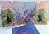 An open vinyl record sleeve and album booklet displayed on a white surface, featuring a surreal illustrated scene with a large bird-like figure, antlered branches, and two small human figures seated in ivy-covered trees. A blue printed insert is placed in front.