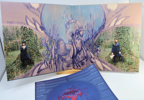 An open vinyl record sleeve and album booklet displayed on a white surface, featuring a surreal illustrated scene with a large bird-like figure, antlered branches, and two small human figures seated in ivy-covered trees. A blue printed insert is placed in front.