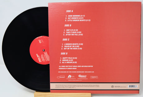 A vinyl record partially pulled from its back cover sleeve, photographed on a wooden stand against a white background. The sleeve is coral red with printed track listings and artist text, while the record is black with a red center label.