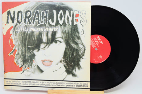 Norah Jones - 'A Little Broken Hearts' vinyl record LP with its illustrated album cover sleeve and a black vinyl record partially visible behind it on a white background.