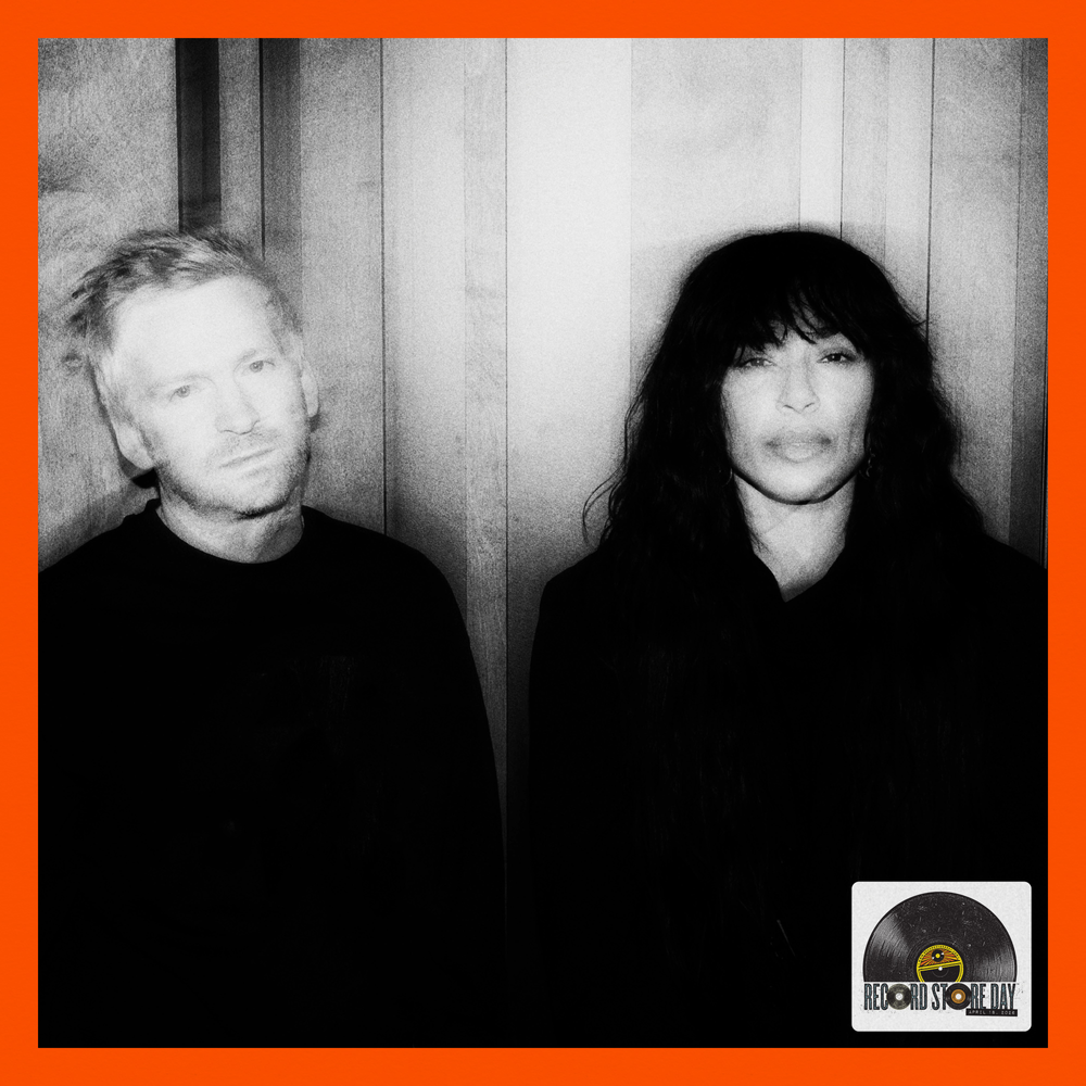 Two people standing against a wooden paneled wall with a record label logo at the bottom right corner.