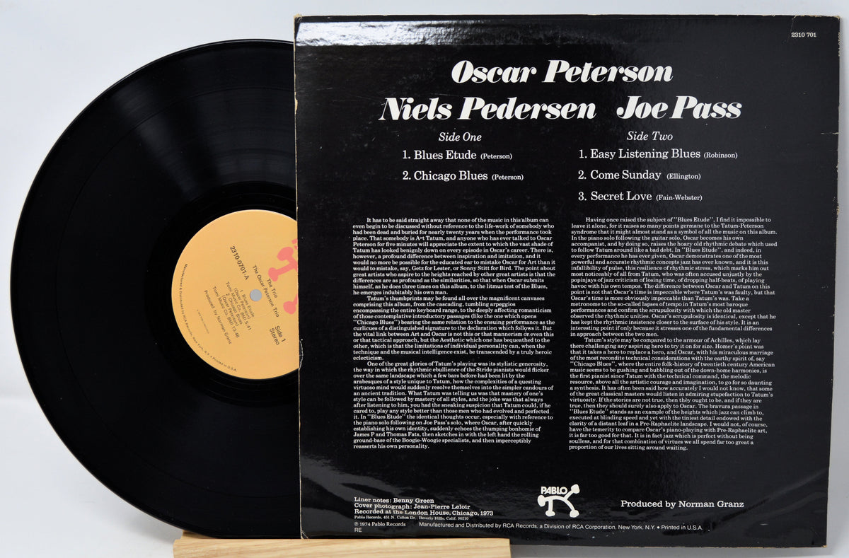 A vintage vinyl record album shown with the black LP partially pulled from its black back cover. The cover reads 'Oscar Peterson Niels Pedersen Joe Pass' and lists track titles and small print.
