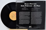 A vintage vinyl record album shown with the black LP partially pulled from its black back cover. The cover reads 'Oscar Peterson Niels Pedersen Joe Pass' and lists track titles and small print.