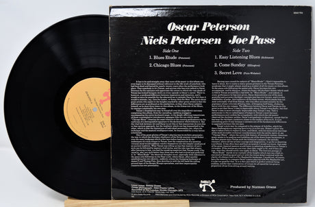 A vintage vinyl record album shown with the black LP partially pulled from its black back cover. The cover reads 'Oscar Peterson Niels Pedersen Joe Pass' and lists track titles and small print.