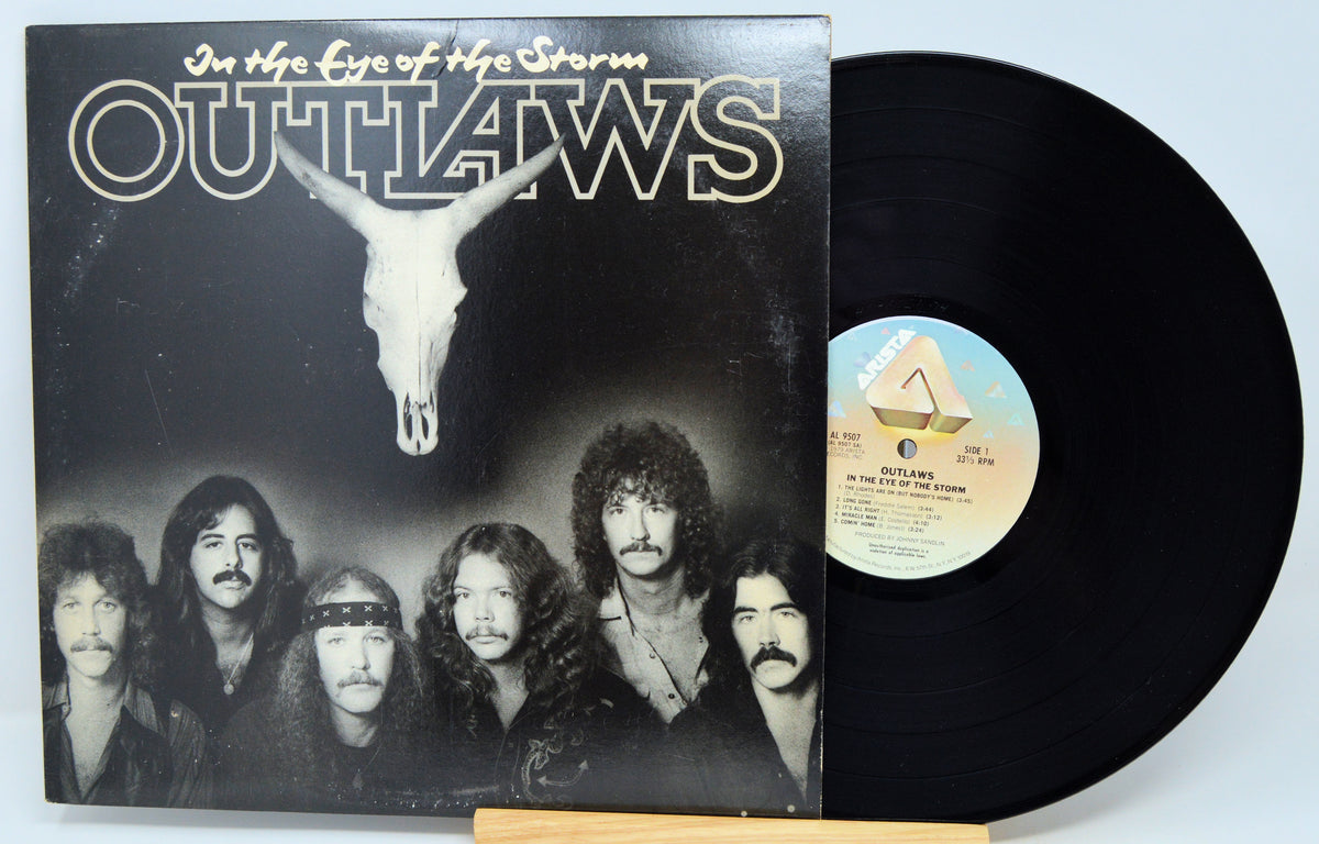 A vinyl record album for 'Outlaws - In the Eye of the Storm' shown with its black LP disc partially pulled from the sleeve, photographed against a plain light background.