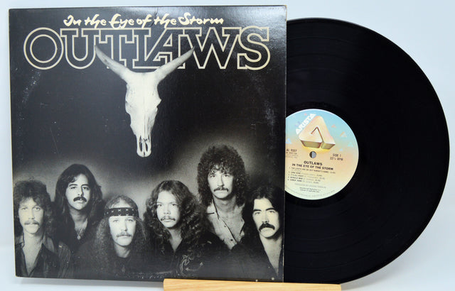 A vinyl record album for 'Outlaws - In the Eye of the Storm' shown with its black LP disc partially pulled from the sleeve, photographed against a plain light background.