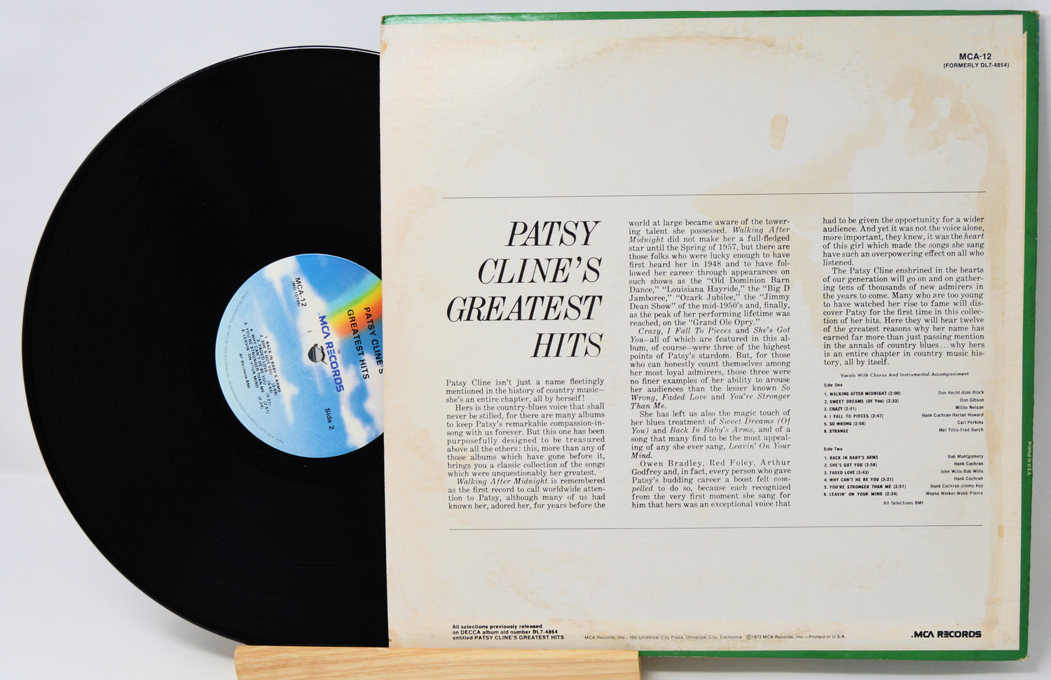 A vintage vinyl record partially pulled from its open sleeve, showing the black record on the left and the cream-colored inner sleeve with printed text on the right. The sleeve has a green edge and shows the title 'Patsy Cline's Greatest Hits.