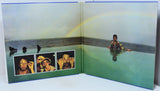 An open book or photo album spread showing a surreal landscape photograph with a rainbow over calm water, a seated figure on the right, three small portrait photos along the lower left, and a blue border around the pages.