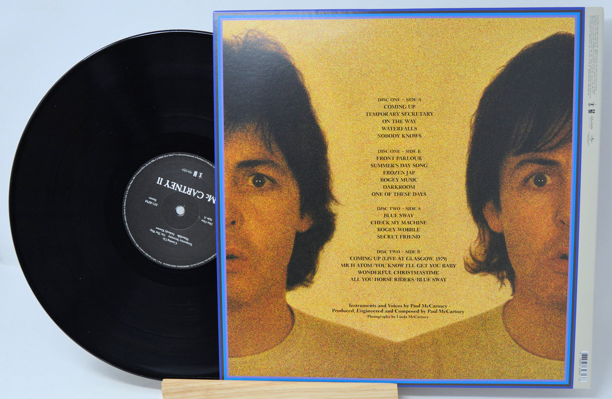 A McCartney II vinyl record partially pulled from its sleeve, showing the back of the album cover with two portraits on a yellow background and tracklist text, alongside the black record on a white surface.