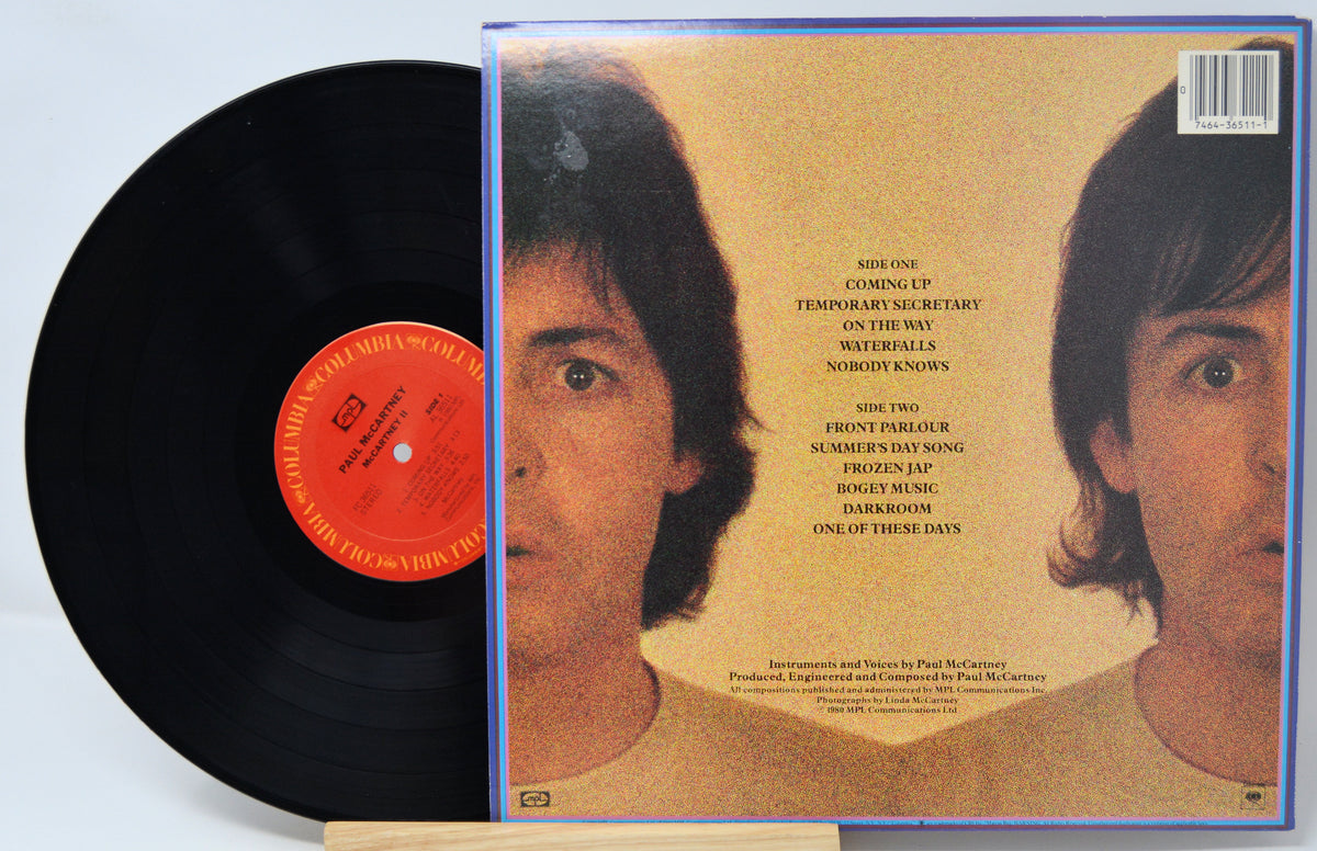 A vinyl record partially pulled from its outer paper sleeve, showing the back of the album cover. The black record has a bright orange center label, and the sleeve features a warm beige background with a close-up portrait of a woman and a track list printed in the center.