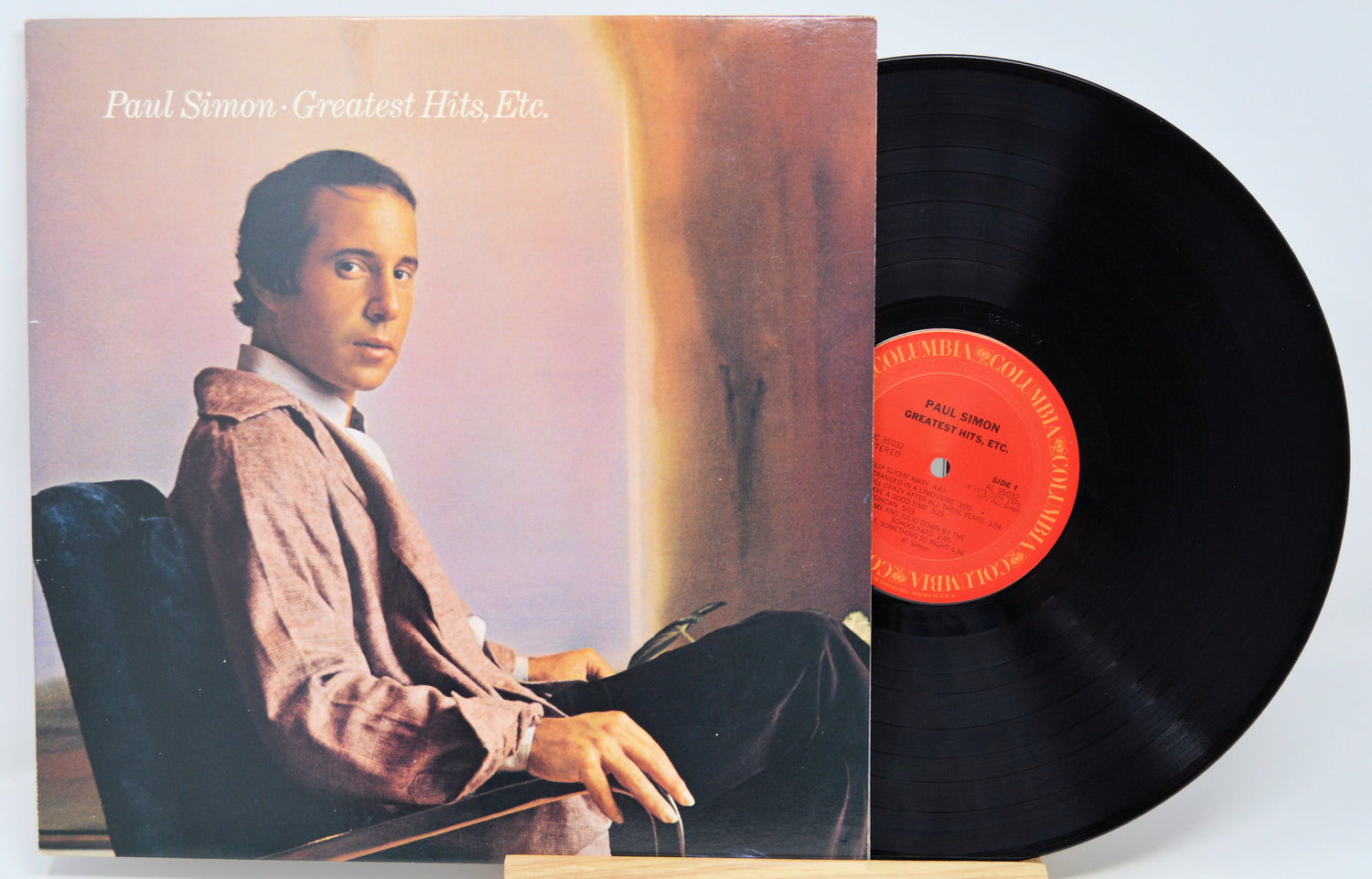 Vinyl record album for Paul Simon's 'Greatest Hits, Etc.' with the album cover standing on the left and the black LP record partially pulled out on the right, photographed against a plain light background.