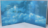 An open vinyl record sleeve or booklet printed with a blue sky and clouds background, displaying song titles in white text on both inner pages.